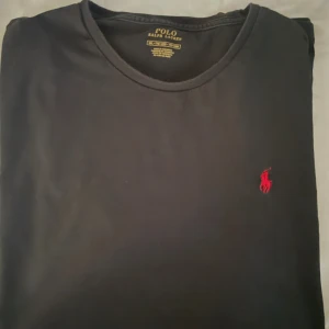 Svart t-shirt från Polo Ralph Lauren - Svart t-shirt från Polo Ralph Lauren i bomull med klassisk rund hals och röd broderad logga på bröstet. Enkel och stilren design som passar perfekt till jeans eller shorts. Skön passform och tidlös look.