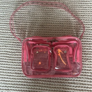 Transparent rosa axelväska från Núnoo - Säljer en transparent rosa axelväska från Núnoo med justerbart axelband och flera fack med dragkedja. Väskan har coola silvriga detaljer och två mindre fickor framtill. Perfekt för dig som vill sticka ut med en unik och trendig väska.Har inte använt den har ingen pris lap när jag köpte den i affären.