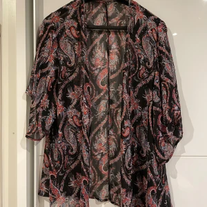 Svart blus med paisleymönster - Säljer en luftig och transparent svart blus med färgstarkt paisleymönster i rött, vitt och blått. 