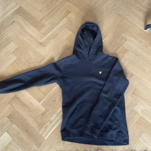 Svart hoodie från Lyle & Scott Junior - Svart hoodie från Lyle & Scott Junior med klassisk gul logga på bröstet. Tröjan har huva, känguruficka och långa ärmar. Perfekt för dig som gillar en enkel och stilren look. Mjuk och skön bomullskänsla.