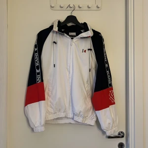 Kani vindjacka i svart, vitt och rött - Snygg vindjacka från Kani i färgblockad stil med svart, vitt och rött. Jackan har huva med snörning, dragkedja framtill och logga på bröstet. Ärmarna har Kani-tryck längs sidorna och elastiska muddar. Perfekt för streetwear-viben.