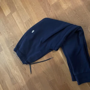 Mörkblå mjukisbyxor Polo Ralph Lauren - Mörkblå mjukisbyxor från Polo Ralph Lauren med klassisk broderad logga på benet. Byxorna har dragsko i midjan och muddar vid bensluten för en skön och avslappnad passform. Perfekta för chill eller streetwear.