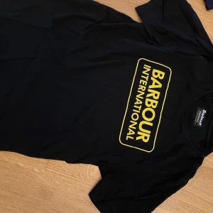 Svart Barbour International t-shirt XXL - Svart t-shirt från Barbour International i storlek XXL med stor gul logga tryckt på bröstet. Klassisk rund hals och korta ärmar. Materialet är mjuk bomull som känns skön mot huden. Perfekt för dig som gillar streetwear och tydliga loggor.