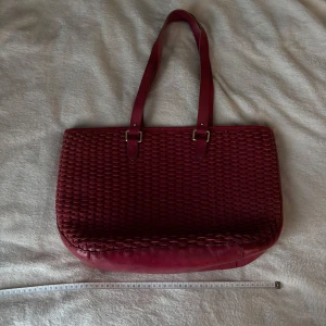 Röd skinnväska från Other Stories - Snygg röd axelväska i flätat skinn från &Other Stories. Väskan har två långa axelband med guldfärgade spännen och ett rymligt huvudfack. Insidan har en ficka med tryckknapp. Ena axelbandet har delat på sig och kan limmas se bild 5.