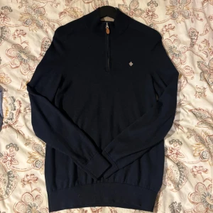 Marinblå merinoulltröja från Morris - Snygg marinblå stickad tröja från Morris i 100% merinoull. Tröjan har half zip med läderdetalj och en liten broderad logga på bröstet. Långärmad med ribbade muddar och hög krage, perfekt för kyliga dagar.