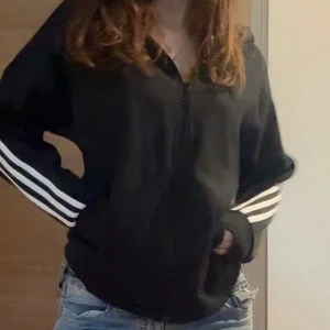 Svart Adidas kofta  - Svart hoodie från Adidas med klassiska vita ränder längs ärmarna och dragkedja framtill. Tröjan har huva och långärmad passform, har även adidas logga vid bröstet. Storlek M. Har en synligt tråd längst ner som är lite uppriven men absolut inget märkbart.