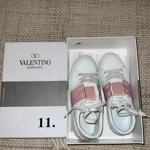 Valentino Garavani Open sneakers vit/rosa - Snygga Valentino Garavani Open sneakers i vitt skinn med bred rosa panel över mitten. Klassisk rund tå, platt sula och vita snören. Lyxig och stilren design med diskreta nitar på sulan. Perfekta för dig som vill sticka ut med exklusiva detaljer.