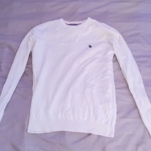 Vit stickad tröja från U.S. Polo Assn. - Vit stickad tröja från U.S. Polo Assn. med rund halsringning och broderad marinblå logga på bröstet. Långärmad modell i mjuk bomull, perfekt för lager-på-lager. Klassisk och stilren look som funkar till allt.