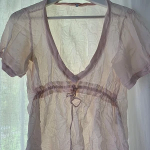 Ljusrosa transparent blus från Gossip - Supersöt ljusrosa transparent blus från Gossip med djup v-ringning och korta ärmar. Blusen har knytband under bysten och tunna kanter i samma färg. Perfekt för lager-på-lager och ger en romantisk vibe.