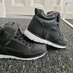 Svarta vinterkängor med snörning - Svarta kängor med vit sula och snörning, fodrade med mjukt material för extra värme. Vattentäta och perfekta för kalla dagar. Sportig design med kardborreband upptill och diskreta sömmar.