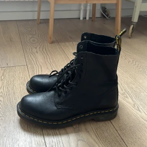 Svarta Dr. Martens boots i skinn - Nästan helt nya klassiska svarta Dr. Martens boots i skinn med rund tå och snörning. Ikonisk gul söm runt sulan och dragflik med logga baktill. Perfekta för dig som vill ha en edgy och tidlös stil.