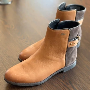 Bruna boots från Lacoste i skinn och mocka - Snygga boots från Lacoste i brunt skinn framtill och mörkbrun mocka baktill. Dekorativt spänne i guld på sidan och rund tå. Perfekta för dig som gillar stilrena detaljer och klassisk design.