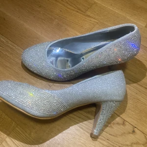 Glittriga silverfärgade pumps - Snygga pumps i silver med massor av glittrande stenar över hela skon. Rund tå och medelhög klack ger en klassisk men ändå festlig look. Perfekta för dig som vill sticka ut och glänsa. Insidan är metallic och skon har en elegant form.