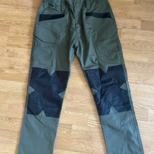 Gröna cargopants Outdoor Tactics M - Säljer ett par gröna cargopants från Outdoor Tactics i storlek M. Byxorna har svarta förstärkningar över knäna i stjärnform, flera stora fickor och justerbara kardborreband på baksidan av benen. Materialet är en blandning av bomull och syntet, perfekt för utomhusaktiviteter.
