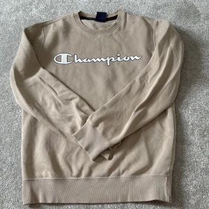 Beige sweatshirt från Champion XS - Snygg beige sweatshirt från Champion i storlek XS. Tröjan har klassisk rund hals och långärmade muddar. Stor Champion-logga i vitt och svart framtill. 