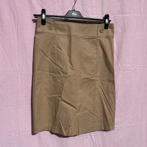 Beige kortkjol från H&M, stl 38 - Snygg beige kjol från H&M i klassisk modell med knappar framtill i midjan och diskret slits bak. Kjolen är tillverkad i mjuk bomull med lite stretch för extra komfort. Perfekt att styla med en enkel topp eller hoodie.