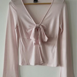 Ljusrosa omlottopp från Gina Tricot - Supersöt ljusrosa topp från Gina Tricot med lång ärm och snygg knytning framtill. Toppen har v-ringning och perfekt för dig som gillar trendig stil.