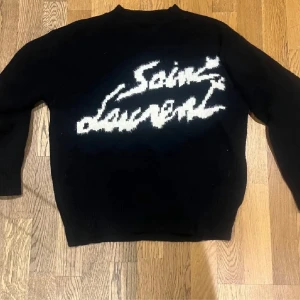 Svart Saint Laurent stickad tröja - Svart stickad tröja från Saint Laurent med vit, stor logga i graffiti-stil över bröstet. Tröjan har lång ärm och klassisk passform, perfekt för dig som vill ha en statement piece med lyxig känsla.