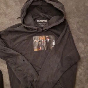 Svart Pulp Fiction hoodie med tryck - Svart hoodie med stor huva och ikoniskt Pulp Fiction-tryck på bröstet samt stor gul logga på ryggen. Mjuk bomullskänsla och klassisk passform. Perfekt för filmälskare som vill sticka ut med en cool streetstyle.