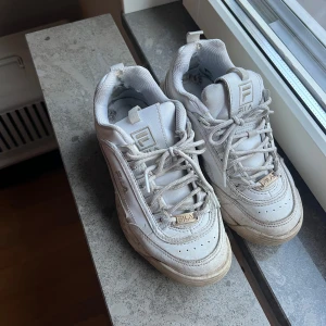 Vita chunky sneakers från Fila - Säljer ett par klassiska vita Fila sneakers med chunky sula och snörning. Skorna har Fila-logga på sidan, tungan och hälen. Ovandelen är i syntet och textil, med detaljer i mesh för extra komfort. Perfekta för dig som gillar streetstyle och sportig look. Slitna i hälen inuti! 