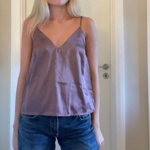 Ljuslila linnetopp med smala axelband - Säljer en ljuslila linnetopp med smala axelband och v-ringad hals. Toppen har en loose passform och är tillverkad i ett glansigt material 