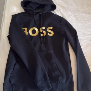 Svart hoodie från BOSS med guldtryck - Svart hoodie från BOSS med stort guldigt BOSS-tryck på bröstet. Klassisk huva med snörning och känguruficka framtill. Tillverkad i mjuk bomullsmix, perfekt för en chill och snygg streetstil. Långärmad och med ribbade muddar för extra komfort. Använd få gånger och i bra skick 