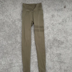 Olivgröna ribbade leggings från Stronger - Snygga olivgröna leggings från Stronger med ribbad struktur och hög midja. De har diskreta ränder på ena benet och logga i midjan. Perfekta för träning eller chill, med tight passform och stretchigt material.