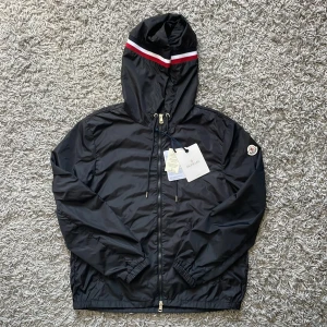 Moncler windbreaker - Svart vindjacka från Moncler med huva och dragkedja. Jackan har Moncler-logga på ärmen och en snygg röd och vit rand på huvan. Tillverkad i lätt polyester, perfekt för blåsiga dagar. Justerbar huva med snörning och elastiska muddar för extra komfort.