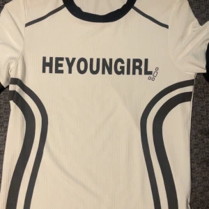 Vit ribbad topp från HEYOUNGGIRL - Snygg vit ribbad topp med svarta detaljer och tryck från HEYOUNGGIRL. Toppen har korta ärmar, svarta grafiska linjer längs sidorna och svart kant vid halsen. Perfekt för dig som gillar sportig och trendig stil.