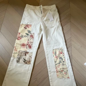 Vita jeans med blommiga patchar - Unika vita jeans med fin passform och blommiga tygpatchar i olika nyanser av rosa och beige på benen. HELT NYA, aldrig andvända. Prislappen kvar. 