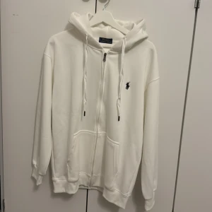Vit hoodie från Polo - Vit hoodie från Polo med dragkedja, huva och snörning. Klassisk svart logga på bröstet och två fickor framtill. Mjuk bomullskänsla och ribbade muddar vid ärmslut och nederkant. Perfekt för chill dagar och streetwear.