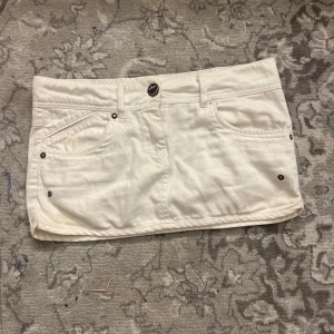 Vit jeanskjol från Xdye, strl 34 - Vit kort jeanskjol från Xdye med klassiska femficksdetaljer, bälteshällor och snygga nitar vid fickorna. Kjolen har knappstängning fram och dekorativa knappar bak. Perfekt för dig som gillar en clean och fräsch look.