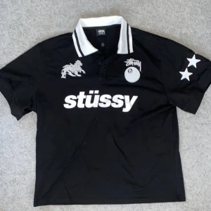 Svart Stüssy t-shirt / piké world tour - Svart t-shirt från Stüssy med vit krage och svarta ränder. Stort Stüssy-tryck på bröstet, broderad lejonlogga och 8-ball detalj. Stjärnor på ena ärmen och stor World Tour-tryck på ryggen. Klassisk streetwear-stil med korta ärmar.