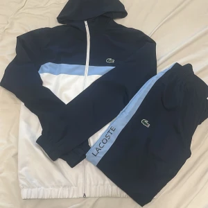 Lacoste vindjacka i blått och vitt - Snygg vindjacka från Lacoste i marinblått, ljusblått och vitt med huva och hel dragkedja. Jackan har sportig design med Lacoste-logga på bröstet och stor SPORT-tryck på ryggen. Perfekt för dig som gillar streetwear och sportig stil.