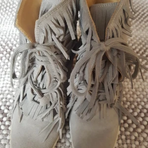 Grå mockaboots med fransar och snörning - Snygga grå boots i mocka med coola fransdetaljer runt skaftet och framtill. Skorna har snörning och platt sula, perfekt för dig som vill sticka ut med din stil. Rund tå och mjukt material gör dem både trendiga och bekväma. Sista bilden är inte på samma skor men typ lika dana ser likadan ut på