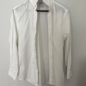 Vit skjorta från T.M. Lewin Regular Fit - Klassisk vit skjorta från T.M. Lewin i regular fit med lång ärm och knappar framtill. Skjortan är tillverkad i bomull och har en stilren, tidlös design med bred krage och manschetter med knappar. Perfekt för dig som gillar en clean och enkel look.