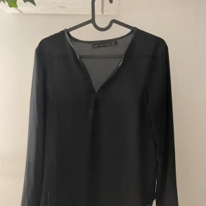 Svart blus med v-ringning från Zara - Snygg svart blus från Zara Basic i tunn, lätt polyester. Blusen har lång ärm och en djup v-ringning som ger en clean och modern vibe. Perfekt att styla med jeans eller kjol för en enkel men chic look.
