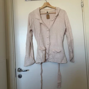 Ljusbeige stickad kofta med knyt - Superfin ljusbeige/ ros stickad kofta från Odd Molly med stora knappar, två fickor framtill och ett knytband i midjan. Koftan har långärmad passform och en söt krage med vågig kant. Perfekt för lager-på-lager och chillare dagar. Storlek 2 som enligt deras storleksguide motsvarar en storlek M.  Mycket fint skick, jag hittar inga synliga anmärkningar eller defekter.