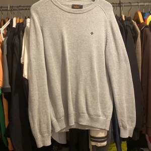 Grå stickad tröja från Sir of Sweden - Snygg ljusgrå stickad tröja från morris med diskret logga broderad på bröstet. Tröjan har rund hals, ribbade muddar och är långärmad. Perfekt för dig som gillar stilrena och klassiska plagg med en modern touch.