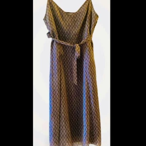 Brun mönstrad klänning Cecilia Classics - Snygg brun klänning från Cecilia Classics med vitt cirkelmönster och knytband i midjan. Klänningen är ärmlös och har en mjuk, flowy passform som ger en chill vibe. Tillverkad i 100% polyester, perfekt för dig som gillar grafiska prints både till fest och vardag! 