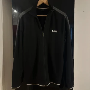 Svart ziptröja från BOSS med grå detaljer - Svart/grå ziptröja från BOSS i storlek M. Tröjan har en liten defekt på ena armen som syns på bilden. Säljs pga den ej passar min stil längre. 