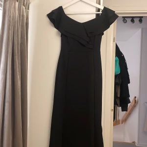 Svart långklänning med volang - Säljer en elegant svart långklänning med offshoulder och bred volang upptill. Klänningen har en stilren och figurnära passform som ger en snygg siluett. Perfekt för dig som vill ha en klassisk look med en modern twist.