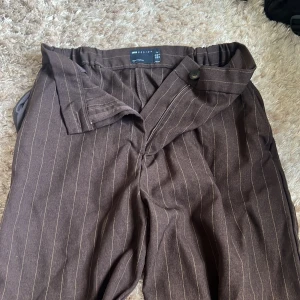 Bruna randiga kostymbyxor Asos Design - Snygga bruna kostymbyxor från Asos Design med kritstrecks ränder. Byxorna har hög midja, elastisk linning bak och klassisk stängning fram med knapp och dragkedja. Benen är vida och modellen är ”tall”. Perfekta för en clean och stilren look.