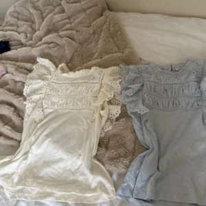 Volangblusar i vitt och ljusblått - Två söta blusar med volangdetaljer och broderade spetsinslag. En i mjuk vit och en i ljusblå färg, båda med korta ärmar och romantisk känsla. Perfekta för dig som gillar feminina och luftiga toppar med extra detaljer. 179 styck. 