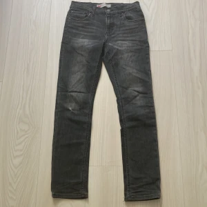 Levi's 512 Slim Taper svarta jeans - Svarta Levi's 512 Slim Taper jeans med snyggt tvättad look och klassiska fem fickor. Jeansen har smal passform med avsmalnande ben och är tillverkade i mjukt denimtyg. Litet hål på framsidan se bild. Label på waist i bak har lossnat i nederkant.