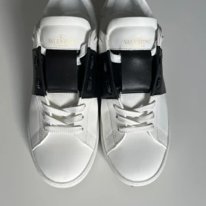 Valentino Garavani Open sneakers vit/svart - Snygga Valentino Open sneakers i vitt skinn med bred svart läderdetalj över snörningen. Klassisk rund tå, vita skosnören och ikoniska nitar baktill på sulan. Perfekta för dig som gillar stilrena och exklusiva sneakers. 