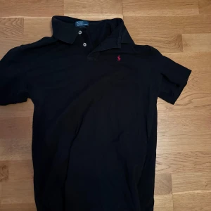 Svart pikétröja från Polo Ralph Lauren - Klassisk svart pikétröja från Polo Ralph Lauren med korta ärmar och röd broderad logga på bröstet. Tröjan har krage och knäppning med två knappar. Perfekt för en clean och stilren look. Xl18-20 passar som en S
