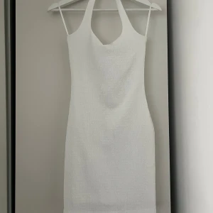 Vit halterneck kortklänning - Säljer en stilren vit kortklänning med halterneck och smala axelband. Klänningen har en tight passform och är tillverkad i ett lätt strukturerat material. Perfekt för sommarens alla tillfällen!