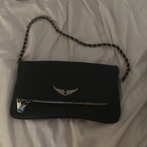 Svart axelväska med kedjerem - Snygg svart axelväska i skinn med silverfärgad kedjerem och dragkedjedetaljer. Framsidan har ett coolt emblem med glittrande vingar. Perfekt storlek för mobilen och det viktigaste. Stilren och edgy design som lyfter vilken outfit som helst. Köpt i Spanien för ca 300kr knappt använd 