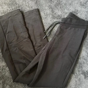 Svarta mjukisbyxor med snörning - Säljer ett par mörkgrå mjukisbyxor med bred resår och snörning i midjan. Byxorna har raka ben och är tillverkade i mjukt bomullsmaterial, perfekta för chill dagar eller när du vill vara extra bekväm.